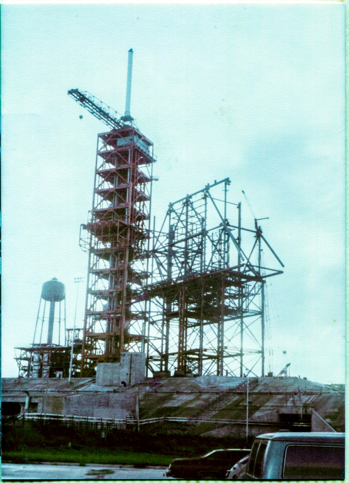 Image 003. This is our very FIRST image of the Pad, taken by James MacLaren on the sly, prior to being issued a camera permit. Viewed from the parking lot just in front of the Sheffield Steel field trailer in the summer of 1980, the the incomplete steel skeleton of the Rotating Service Structure at Space Shuttle Launch Complex 39-B, Kennedy Space Center, Florida, stands supported on its temporary falsework support framing, growing into the sky, as it is assembled by the Union Ironworkers of Local 808, working for Wilhoit Steel Erectors. To its immediate left, stands the taller red framework of the Fixed Service Structure, topped by the Hammerhead Crane, and the Lightning Mast above that. Photo by James MacLaren.